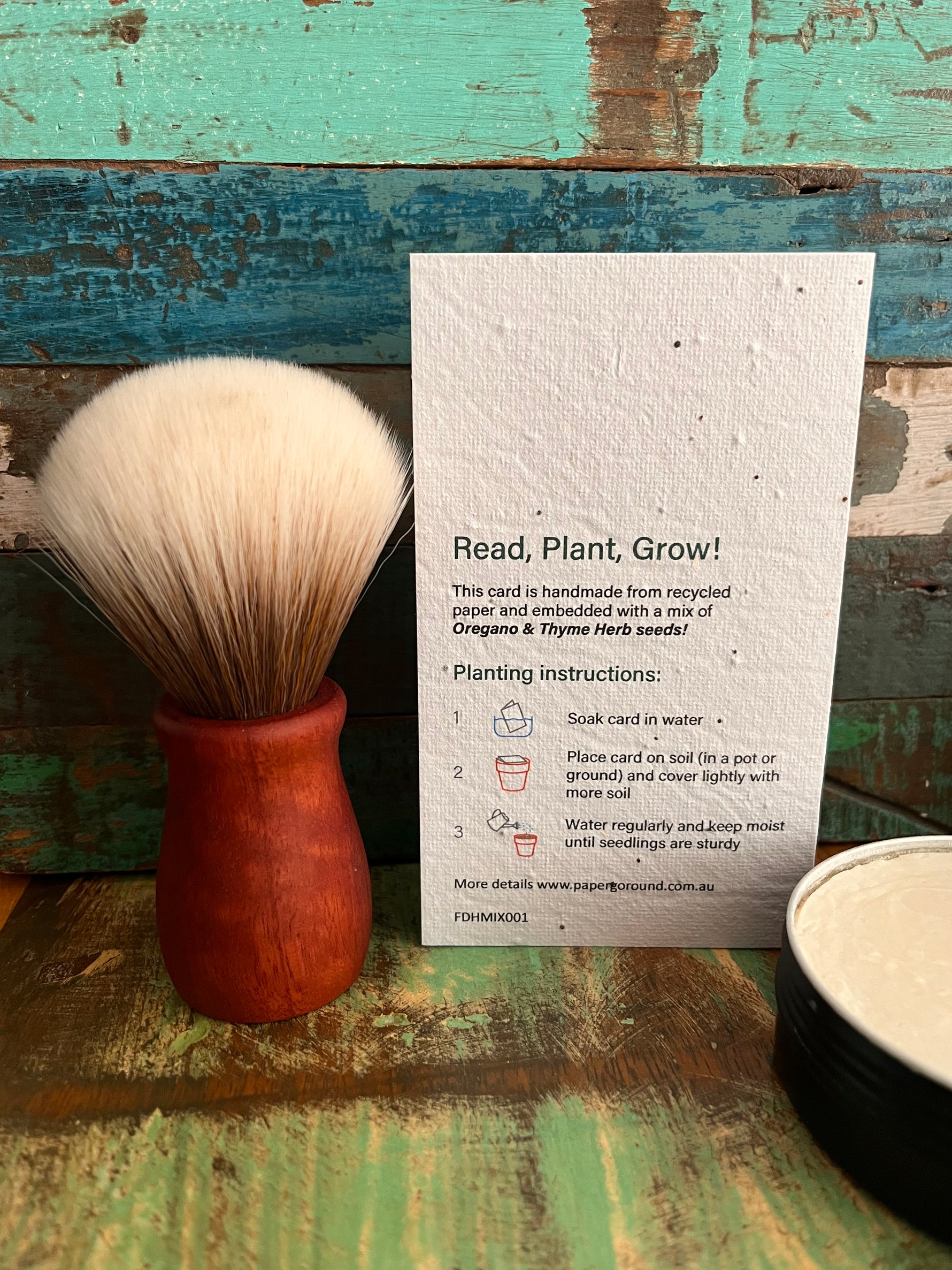 A handmade seed paper card with planting instructions stands upright on a rustic painted wooden surface next to a wooden-handled shaving brush with cream bristles and a round tin of white product.