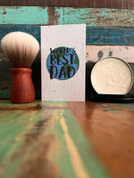 A handmade paper greeting card reading 'WORLD'S BEST DAD' with a blue and green globe graphic, styled on a rustic wooden surface next to a shaving brush and a tin of shaving soap.