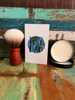 A handmade greeting card reading 'World's Best Dad' in a blue and green circular design, styled on a rustic painted wooden surface alongside a shaving brush and a tin of shaving soap.