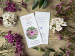 Plantable memorial bookmarks laid flat on a wooden surface, surrounded by white and purple flowers and green leaves. 