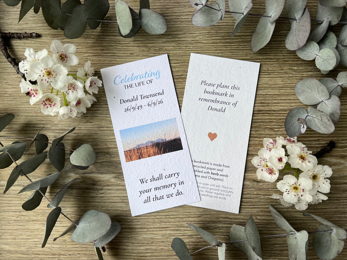 Seed paper printed memorial bookmarks laid flat on a wooden surface, surrounded by eucalyptus leaves and small white flowers. 