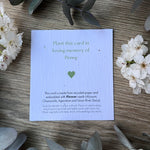 A plantable memorial seed card with green text and a small green heart, placed on a wooden surface with white flowers around it. The card reads, 'Plant this card in loving memory of Penny' and includes planting instructions.