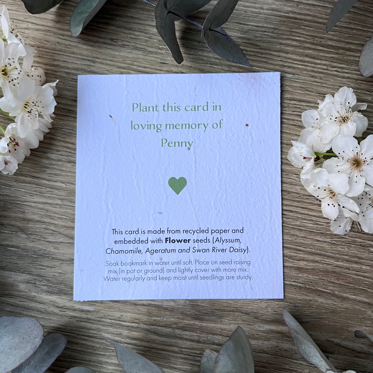 A plantable memorial seed card with green text and a small green heart, placed on a wooden surface with white flowers around it. The card reads, 'Plant this card in loving memory of Penny' and includes planting instructions.