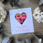 A seed paper memorial card with a heart-shaped floral photo. The card reads 'In Loving Memory,' 'Penny Anderson,' dates, and 'Forever Cherished.