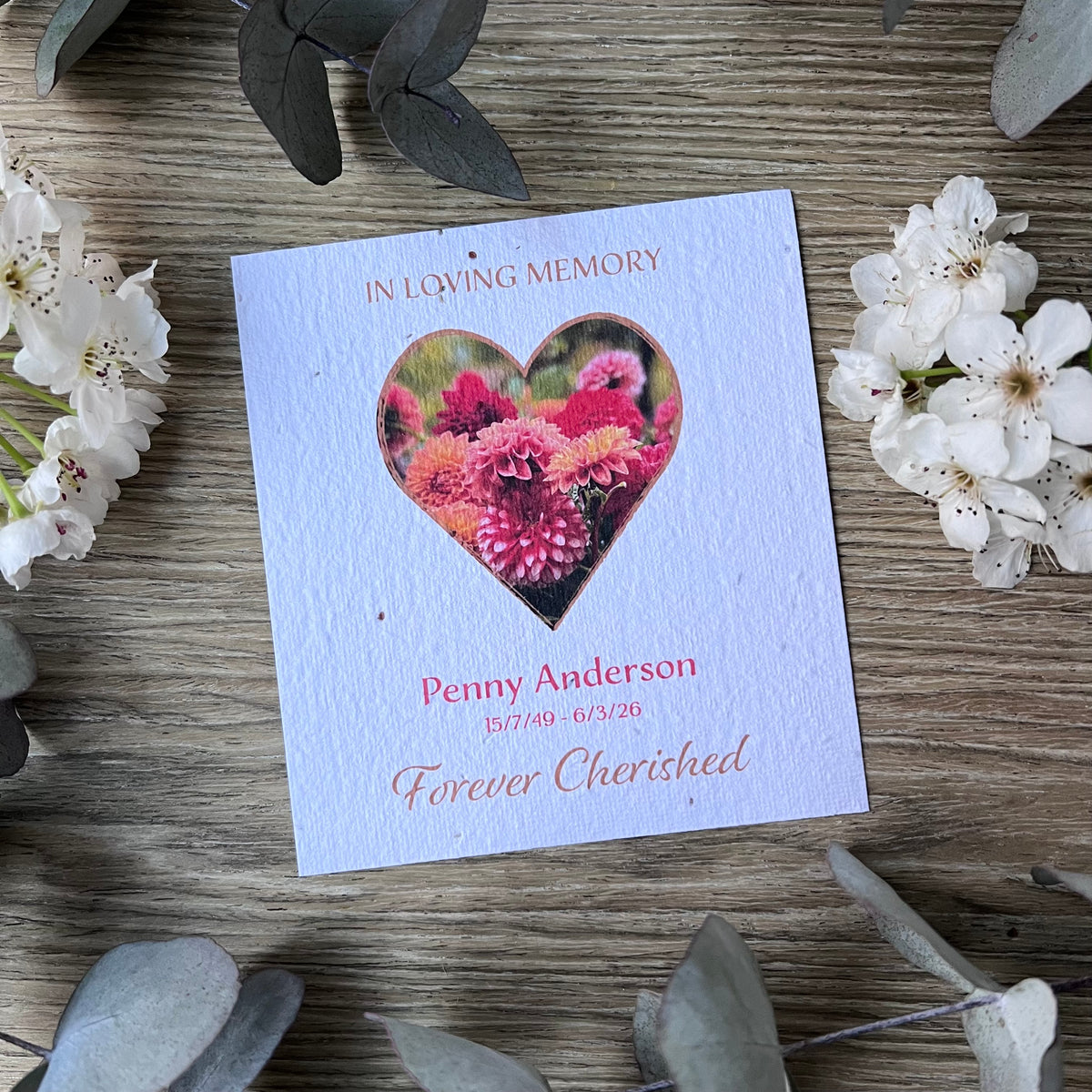 A seed paper memorial card with a heart-shaped floral photo. The card reads 'In Loving Memory,' 'Penny Anderson,' dates, and 'Forever Cherished.