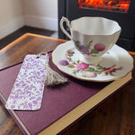 Teacup with floral design on a saucer, seed paper bookmark, and book on a wooden surface.