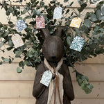 Display rabbit figure with a beige scarf surrounded by eucalyptus leaves and decorative tags on a wooden background.