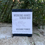 A rectangular bar of Working Hands Scrub Bar labeled 'Volcanic Pumice' stands upright outdoors on a stone surface with green foliage in the background.