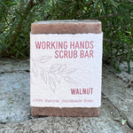 A rectangular bar of walnut hand scrub soap wrapped in a textured white paper sleeve with pink text, set against a background of green foliage and concrete.